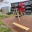 Bench, Steel Construction na dětském hřišti s houpačkovou sestavou a obytnými budovami v pozadí.