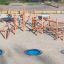 Sonora dřevěná lezecká konstrukce se třemi modrými zapuštěnými trampolínami na pískovém hřišti