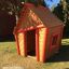 Dřevěný domeček na hraní pro děti - Crooked House, hnědý s červeným lemováním, venkovní prostředí.