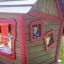 Dřevěný herní domek s červeným lemováním a usmívajícím se dítětem vykukujícím z okna z Anna's Playhouse