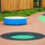 Round trampoline (100 cm) zapuštěný do povrchu hřiště se zelenou skákací podložkou a černým okrajem.