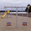 Baby Double Swing na hřišti s pískovým povrchem a žlutou skluzavkou v pozadí.