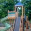 Twister castle with slide na hřišti s pískovým podkladem a zelení v pozadí.